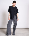 A person stands indoors against a plain background, wearing the jan-n-june SLITTED T-SHIRT NADIL BLACK styled with wide-leg gray jeans, looking down with hands relaxed by their sides.