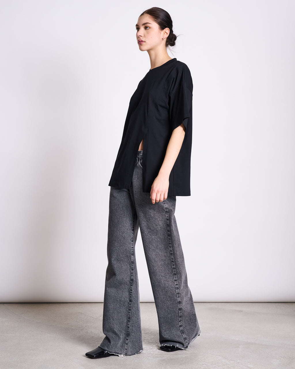 A person stands sideways against a plain background, wearing the jan-n-june SLITTED T-SHIRT NADIL BLACK, wide-leg gray jeans, and black shoes. Their hair is pulled back and they have a neutral expression.