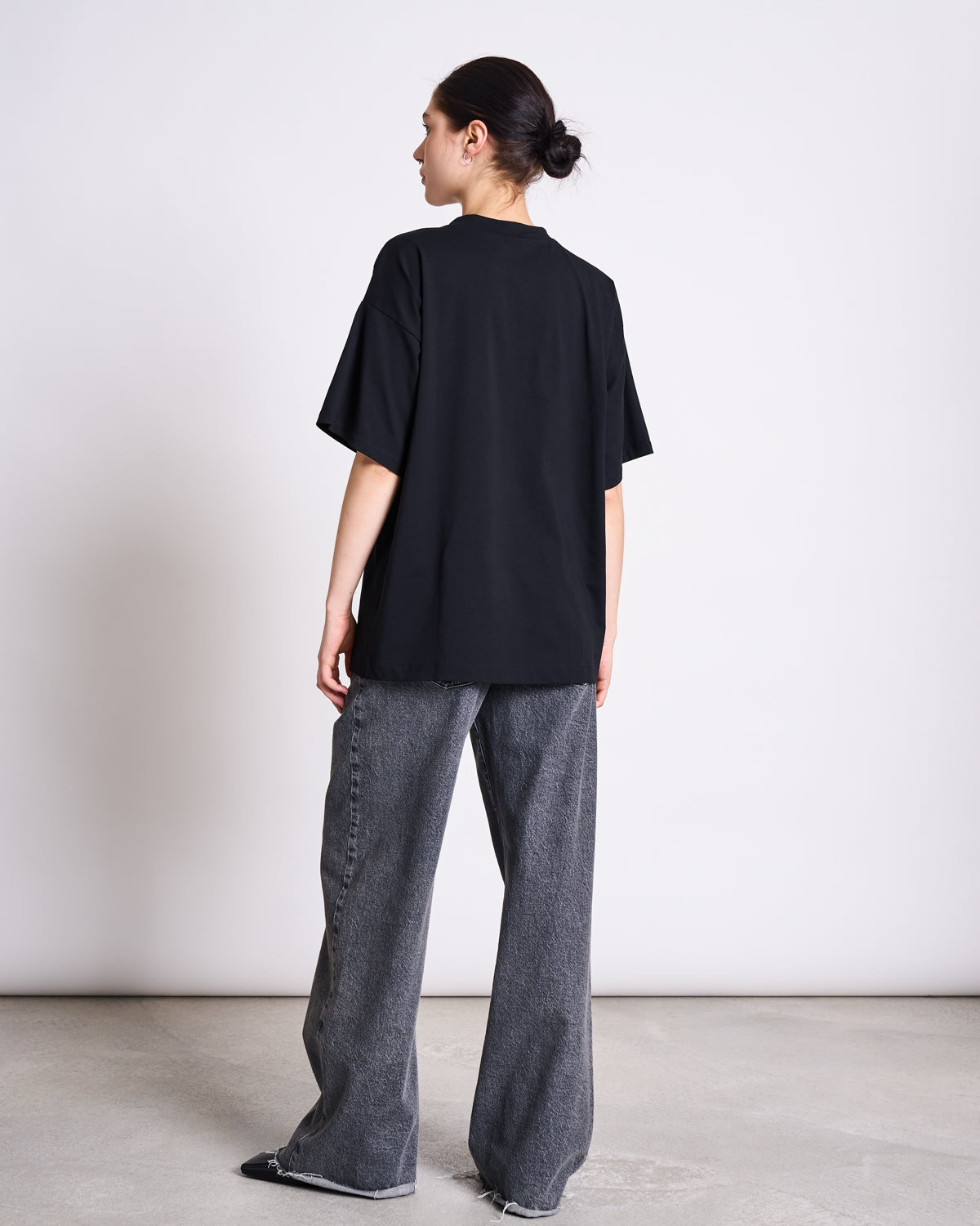 A person with dark hair in a low bun stands facing away, wearing the jan-n-june SLITTED T-SHIRT NADIL BLACK and wide-leg gray jeans with frayed hems, set against a plain light background.