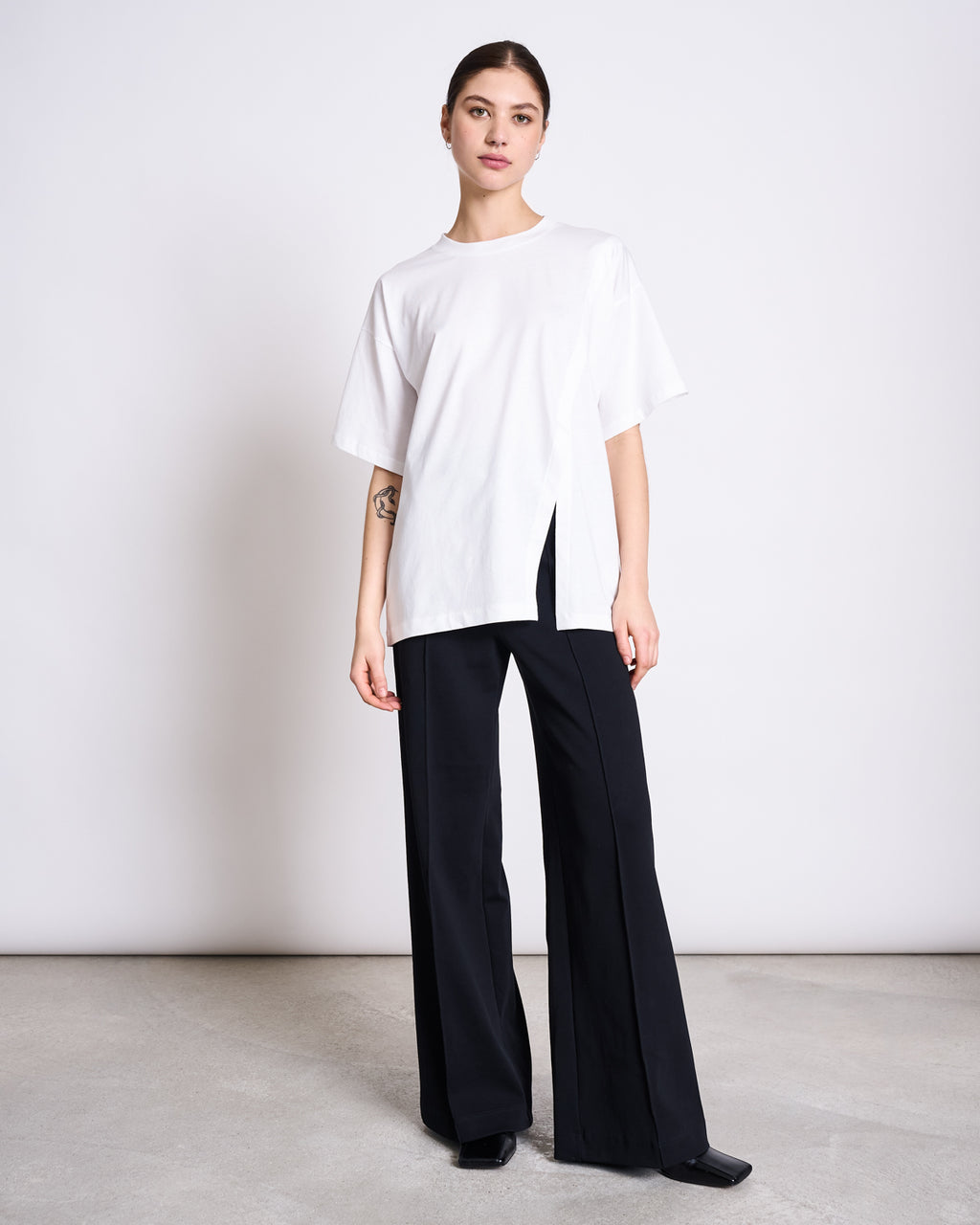 A woman stands against a plain background wearing the jan-n-june SLITTED T-SHIRT NADIL WHITE—an oversized, recycled organic cotton tee with a side slit—paired with wide-leg black pants and black shoes.