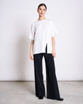 A woman stands against a plain background wearing the jan-n-june SLITTED T-SHIRT NADIL WHITE—an oversized, recycled organic cotton tee with a side slit—paired with wide-leg black pants and black shoes.