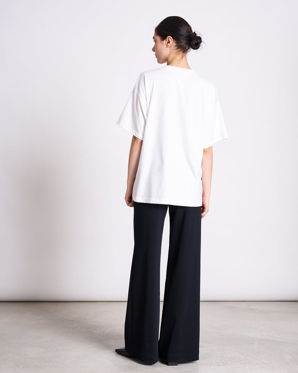 A person with dark hair in a low bun faces away, wearing the jan-n-june SLITTED T-SHIRT NADIL WHITE, an oversized tee made from recycled organic cotton, and wide-leg black pants against a plain white background.