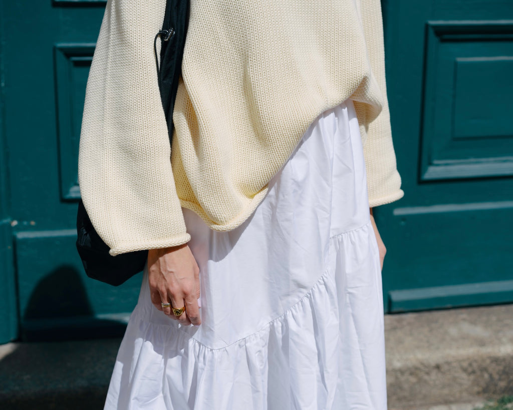 A person wearing the jan-n-june JUMPER SONA BUTTERMILK GOTS stands before a teal door, paired with a white tiered skirt. Visible are only their midsection and hand, showcasing gold rings and a black bag.