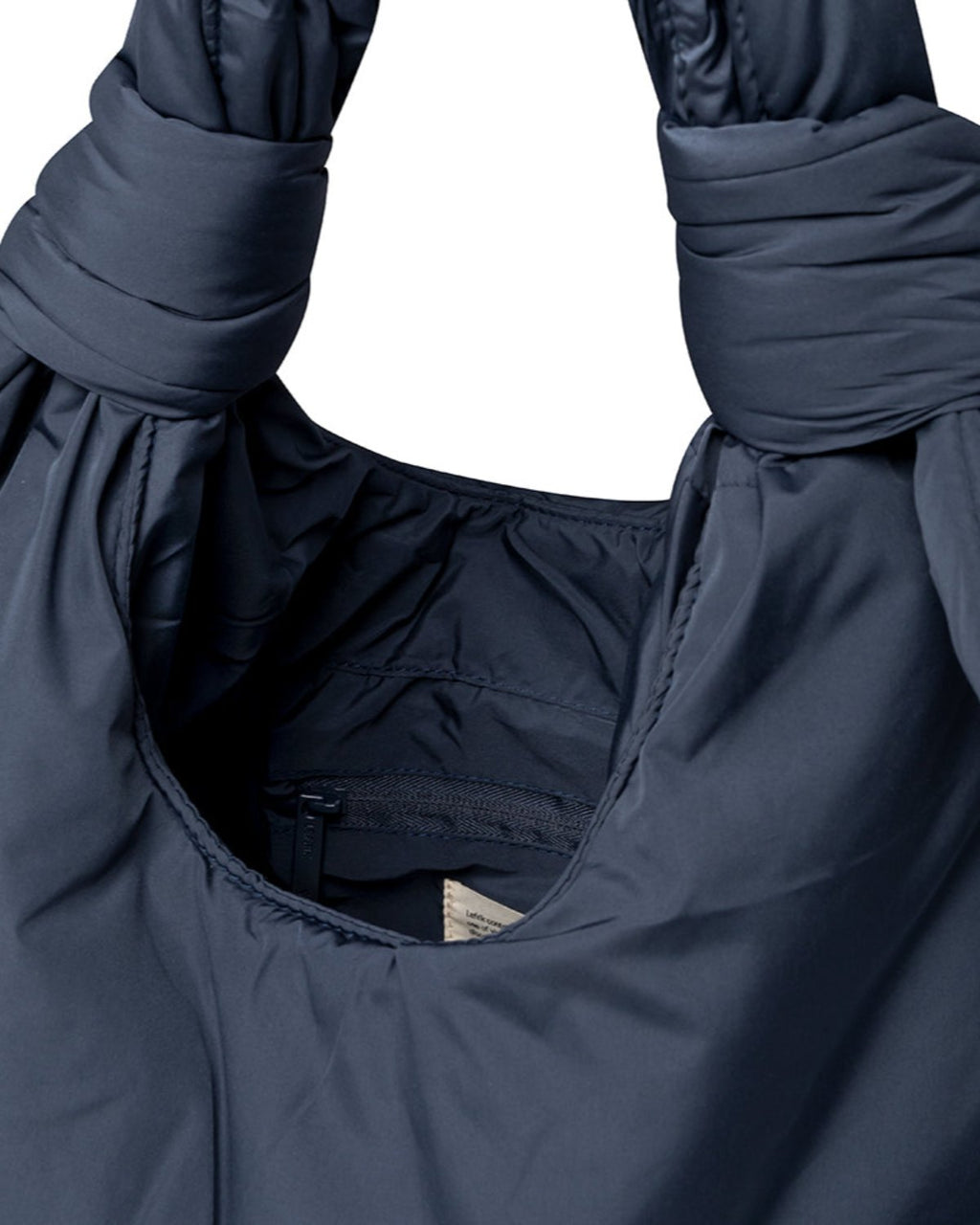 Close-up of the jan-n-june BAG BIWA PUFFY REGULAR MAIBON, a dark blue quilted bag with ruched handles and visible interior stitching. Made from recycled polyester, it has a small white tag partially showing inside.