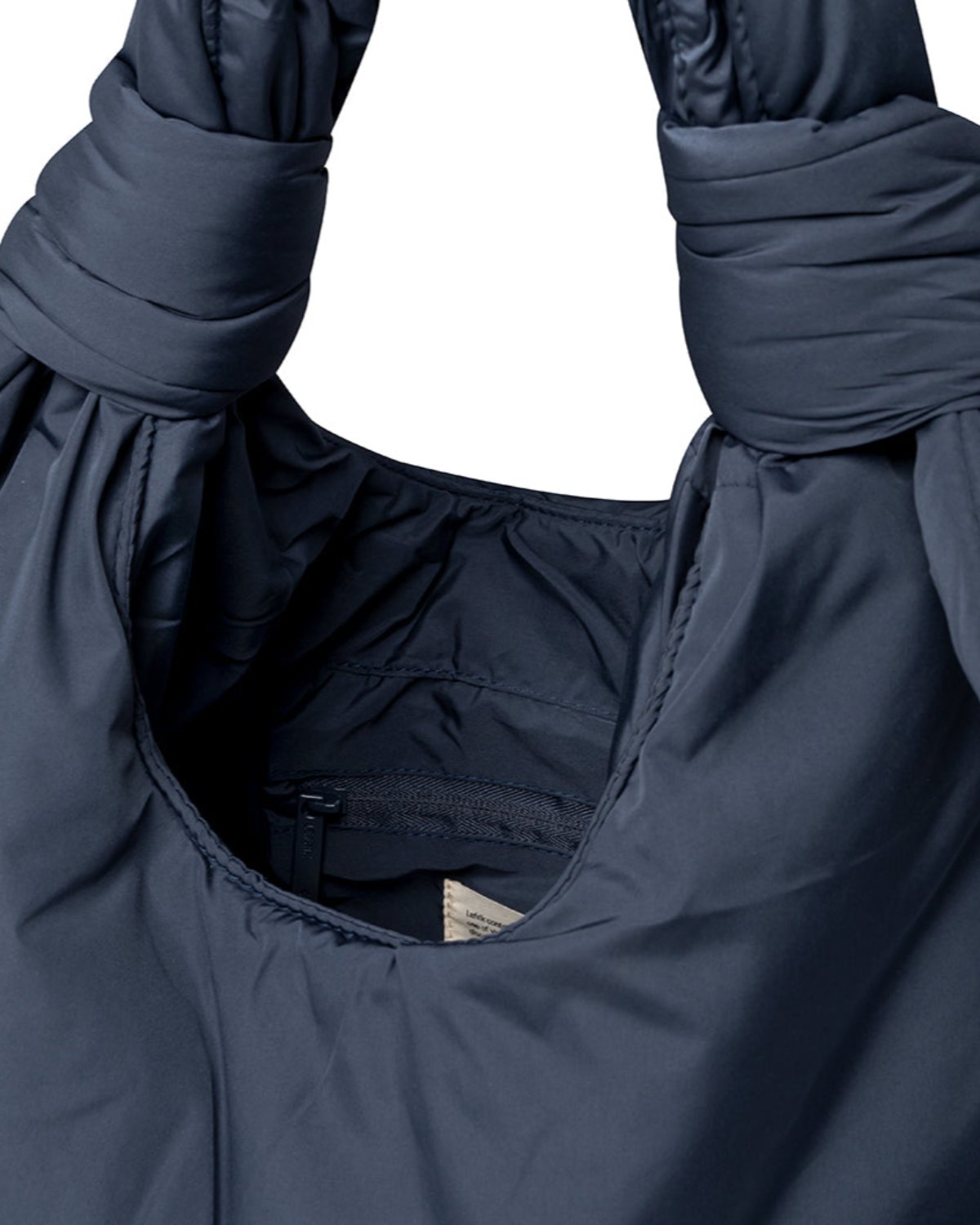 Close-up of the jan-n-june BAG BIWA PUFFY REGULAR MAIBON, a dark blue quilted bag with ruched handles and visible interior stitching. Made from recycled polyester, it has a small white tag partially showing inside.