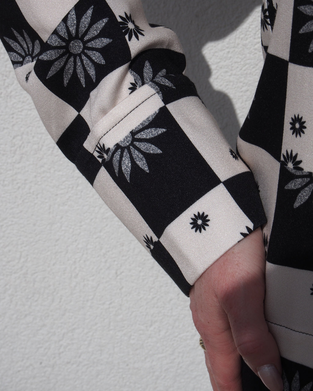 A close-up of a person’s arm and hand in the jan-n-june TIE BLOUSE KALUZ FLORAL SQUARE PRINT, featuring a black and cream floral checkered design, standing against a light-colored textured wall.