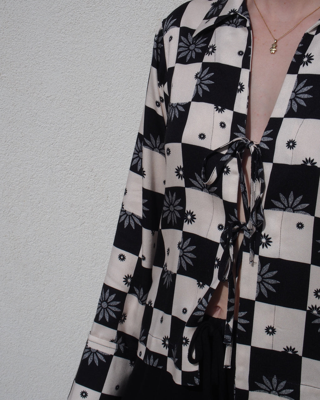 A person wearing the jan-n-june TIE BLOUSE KALUZ FLORAL SQUARE PRINT, featuring black and white checks with floral accents and chest ties, stands by a light textured wall, accessorized with a gold necklace.