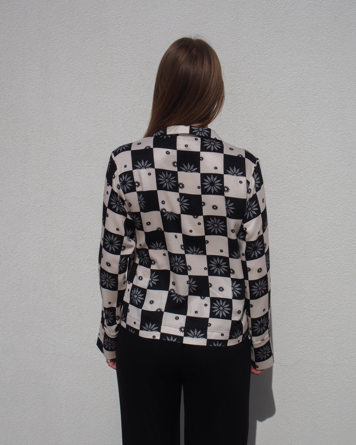 A person with long brown hair stands facing a light wall, wearing a black and white jan-n-june TIE BLOUSE KALUZ FLORAL SQUARE PRINT and black pants.