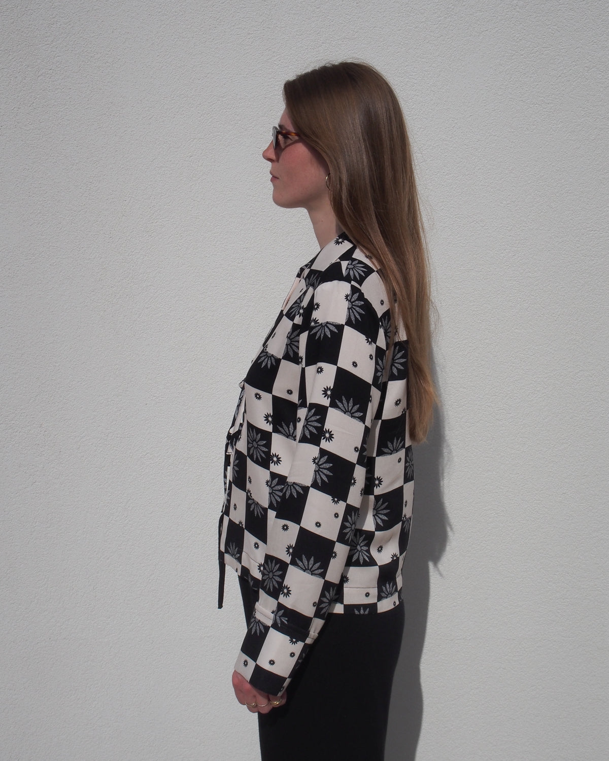 A woman with long brown hair and sunglasses stands in profile against a white wall, wearing the jan-n-june TIE BLOUSE KALUZ FLORAL SQUARE PRINT and black pants.