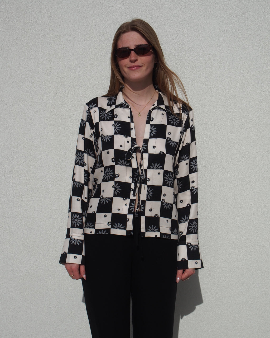 A woman with long hair and sunglasses stands against a white wall, facing forward in a relaxed pose, wearing jan-n-junes TIE BLOUSE KALUZ FLORAL SQUARE PRINT with black pants.