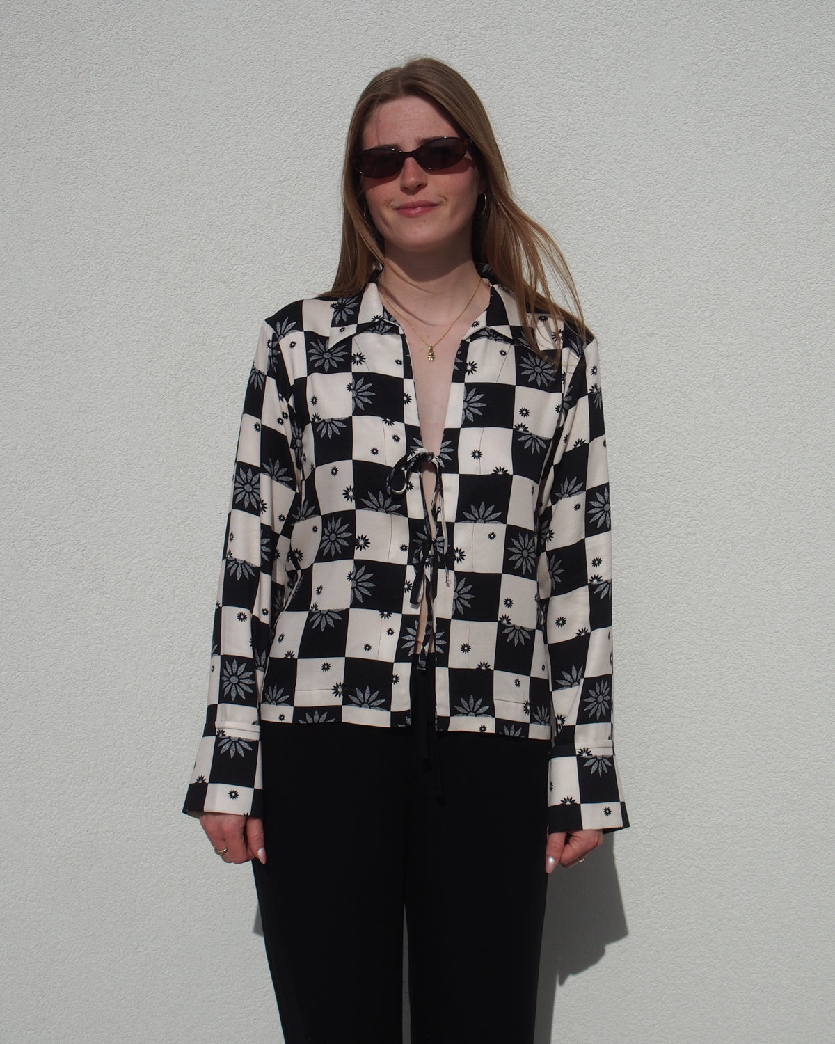 A woman with long hair and sunglasses stands against a white wall, facing forward in a relaxed pose, wearing jan-n-junes TIE BLOUSE KALUZ FLORAL SQUARE PRINT with black pants.