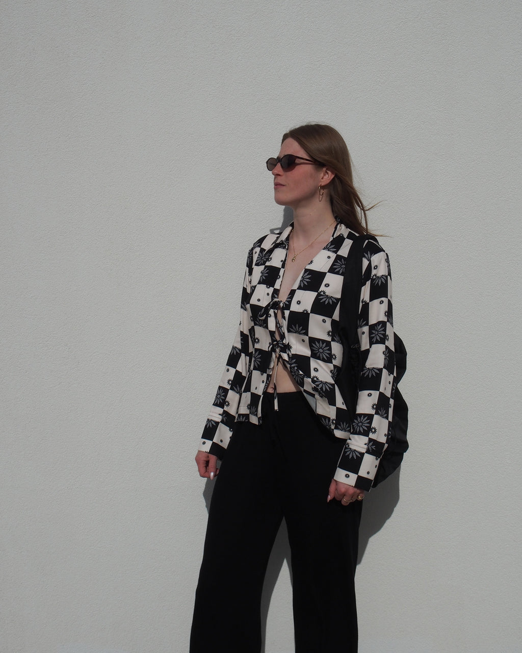 A woman in sunglasses and the jan-n-june TIE BLOUSE KALUZ FLORAL SQUARE PRINT, paired with black pants, stands against a white wall, looking to the side with her hands relaxed by her sides.