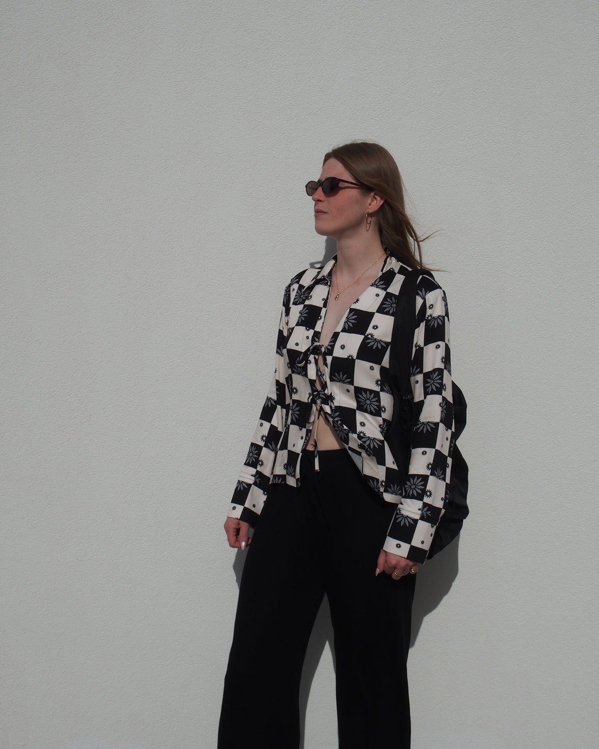 A woman in sunglasses and the jan-n-june TIE BLOUSE KALUZ FLORAL SQUARE PRINT, paired with black pants, stands against a white wall, looking to the side with her hands relaxed by her sides.