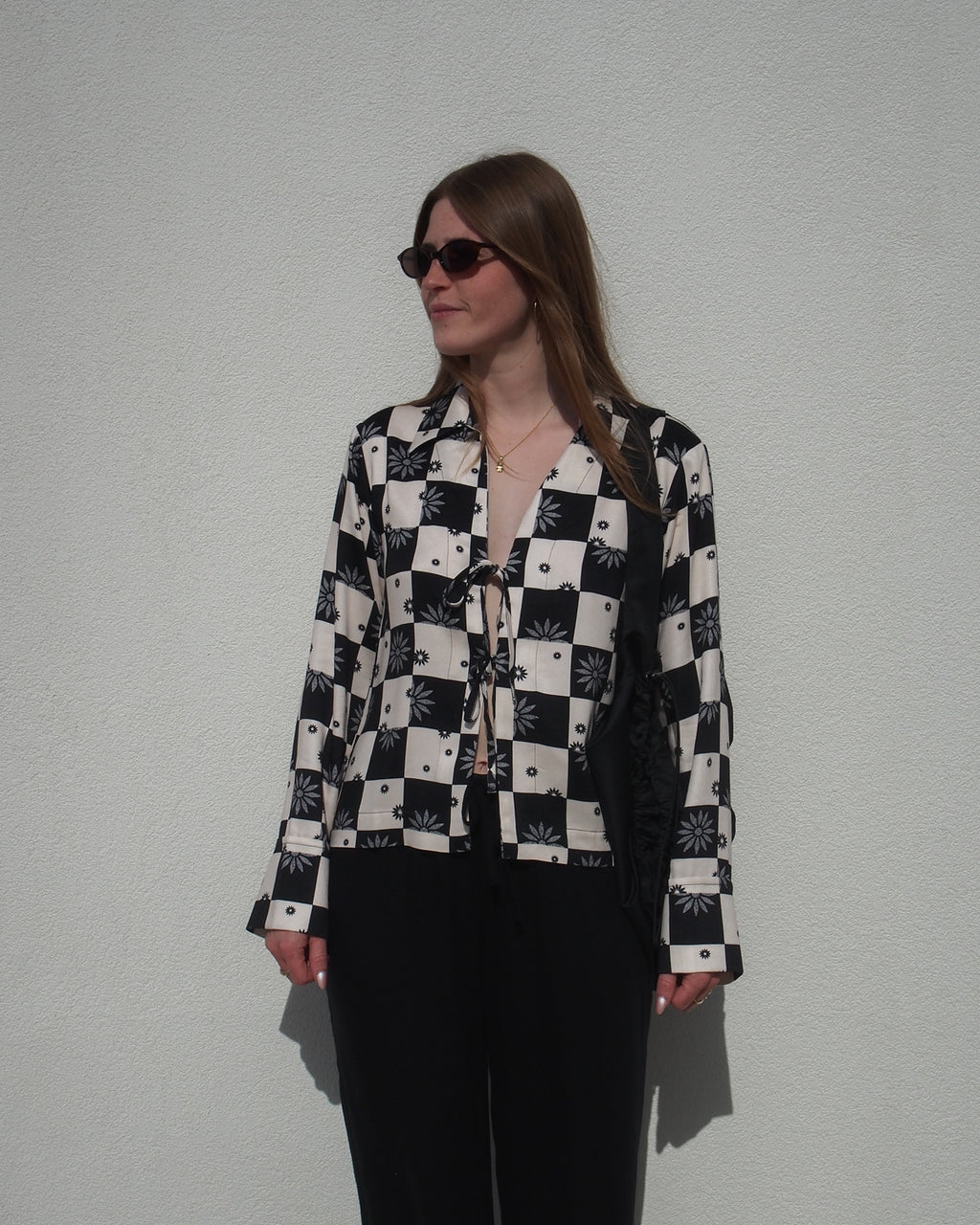 A woman in sunglasses wears the jan-n-june TIE BLOUSE KALUZ FLORAL SQUARE PRINT, featuring a black-and-white checkered and floral design, as she stands relaxed against a plain white wall.