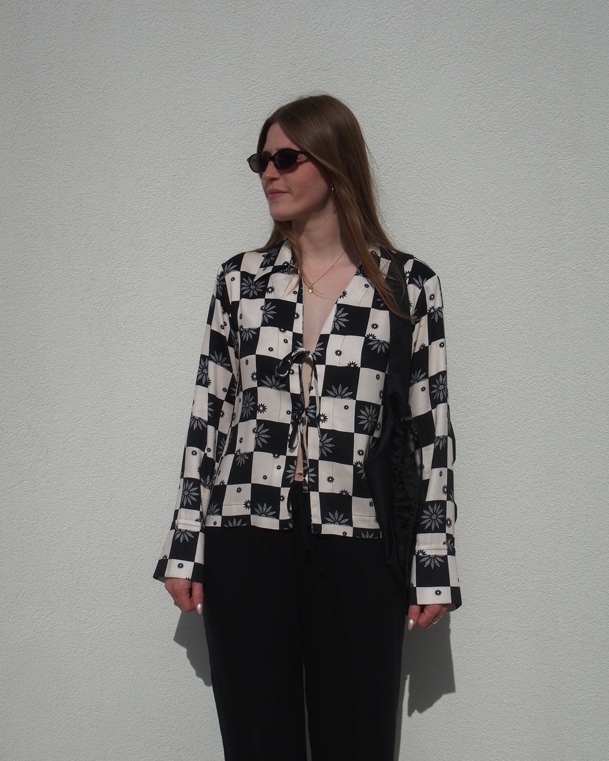 A woman in sunglasses wears the jan-n-june TIE BLOUSE KALUZ FLORAL SQUARE PRINT, featuring a black-and-white checkered and floral design, as she stands relaxed against a plain white wall.