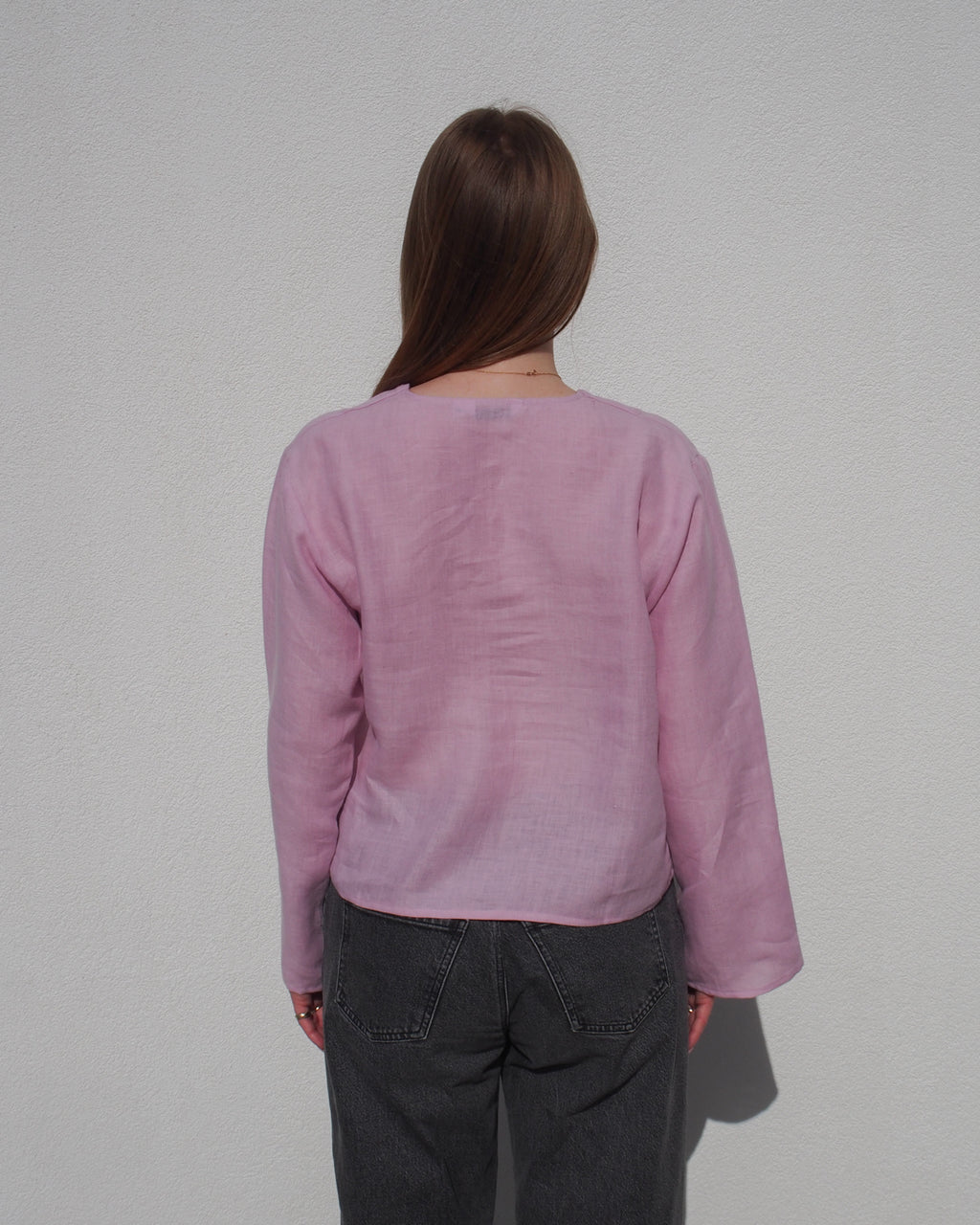 A person with long brown hair, wearing the jan-n-june LINEN TIE BLOUSE SKAGEN LIGHT ORCHID, stands in dark gray jeans facing a plain white wall with their back to the camera.