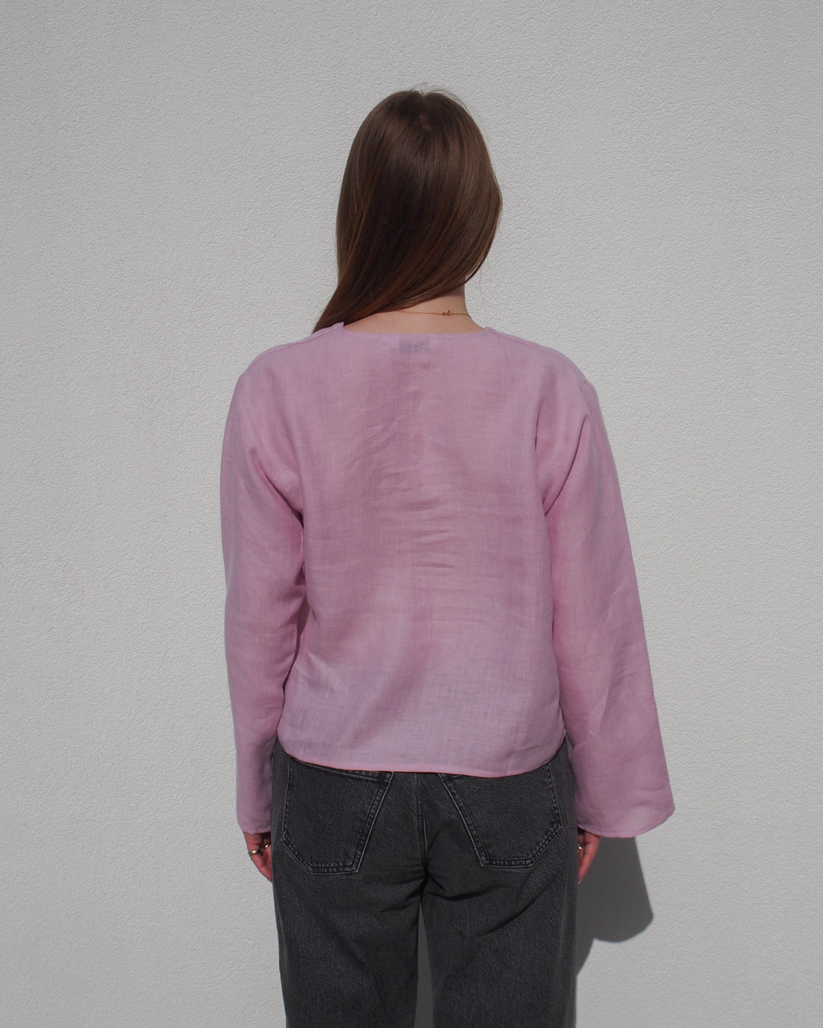 A person with long brown hair, wearing the jan-n-june LINEN TIE BLOUSE SKAGEN LIGHT ORCHID, stands in dark gray jeans facing a plain white wall with their back to the camera.