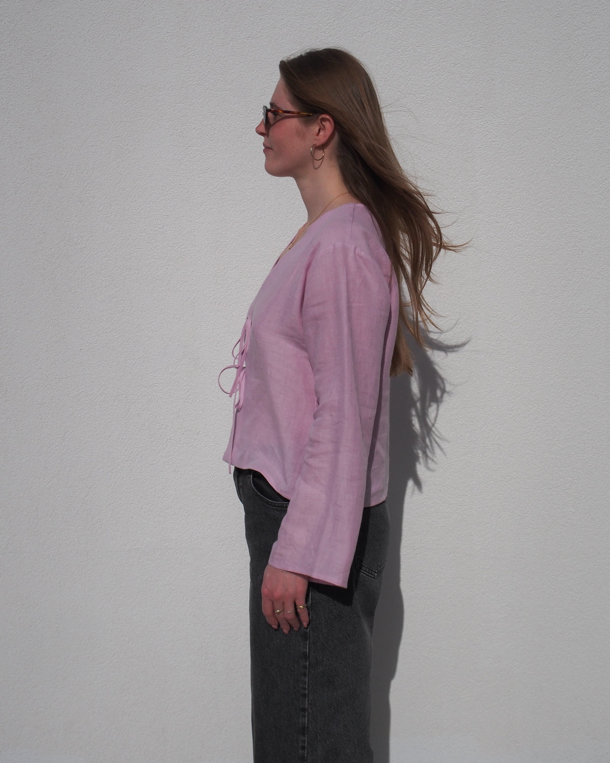 A woman with long brown hair stands in profile against a white wall, wearing the jan-n-june LINEN TIE BLOUSE SKAGEN LIGHT ORCHID, dark jeans, and sunglasses. Her hair is gently blowing in the wind.