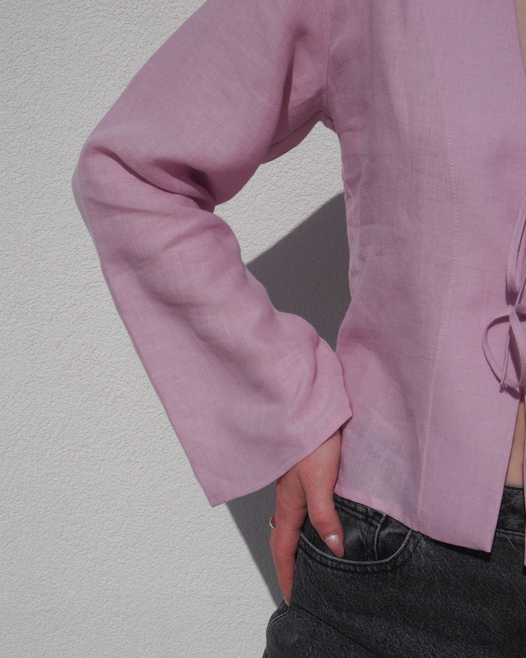 A person stands against a white wall wearing the jan-n-june LINEN TIE BLOUSE SKAGEN LIGHT ORCHID with dark jeans, one hand on their hip. Only their torso and part of the left arm are visible.