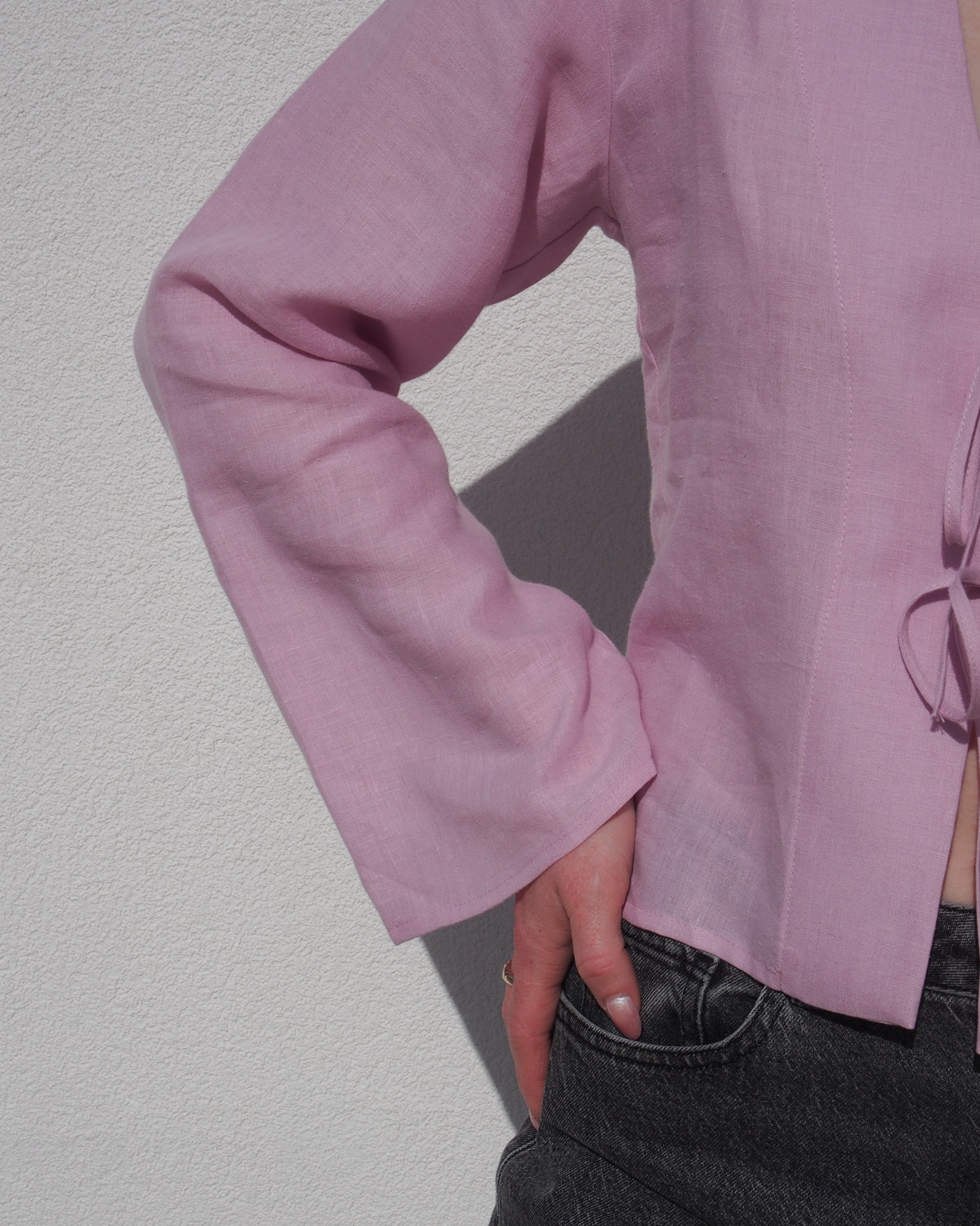 A person stands against a white wall wearing the jan-n-june LINEN TIE BLOUSE SKAGEN LIGHT ORCHID with dark jeans, one hand on their hip. Only their torso and part of the left arm are visible.