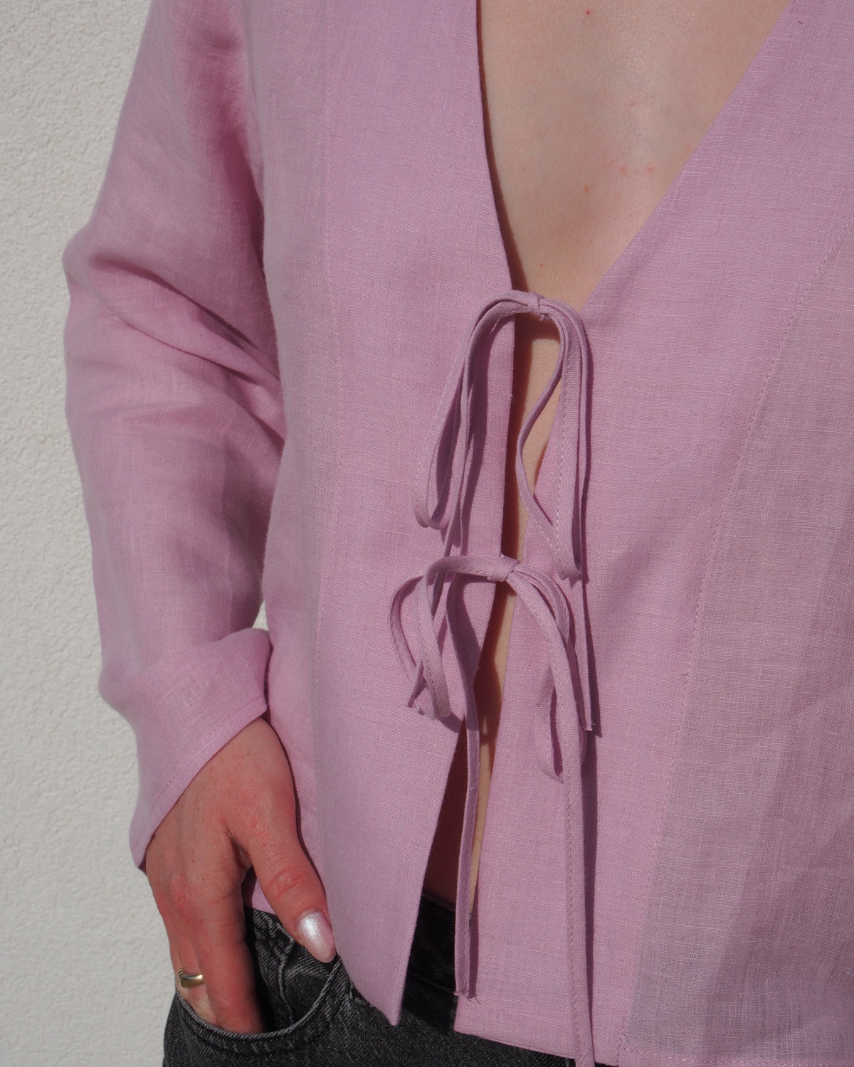 Wearing the jan-n-june LINEN TIE BLOUSE SKAGEN LIGHT ORCHID with a deep V neckline and black denim jeans, a person rests their left hand in their pocket while standing by a light-colored wall.
