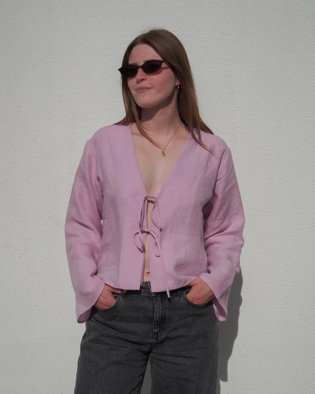 A person wearing the jan-n-june LINEN TIE BLOUSE SKAGEN LIGHT ORCHID, paired with dark sunglasses and jeans, stands against a plain white wall with hands in pockets.