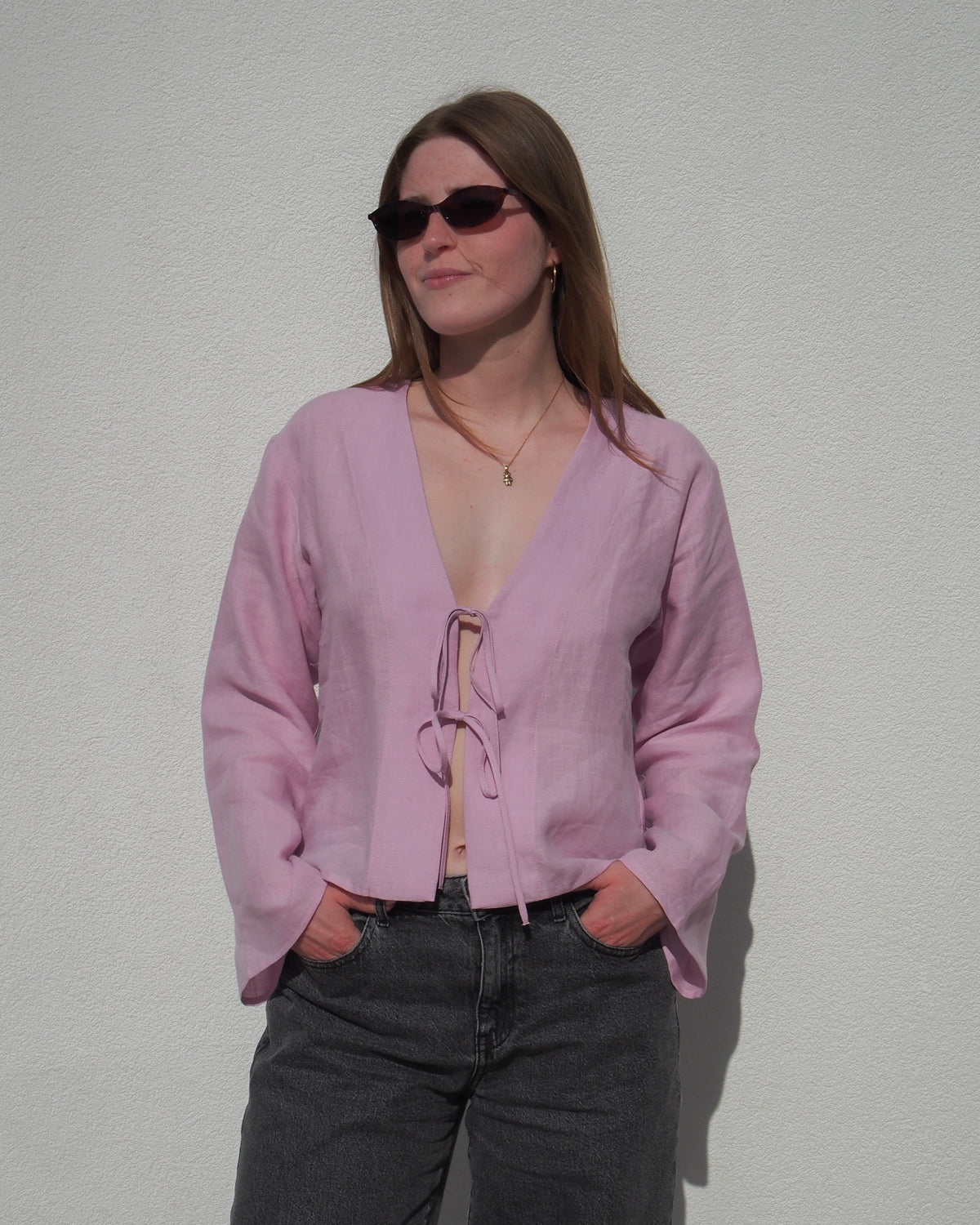 A person wearing the jan-n-june LINEN TIE BLOUSE SKAGEN LIGHT ORCHID, paired with dark sunglasses and jeans, stands against a plain white wall with hands in pockets.