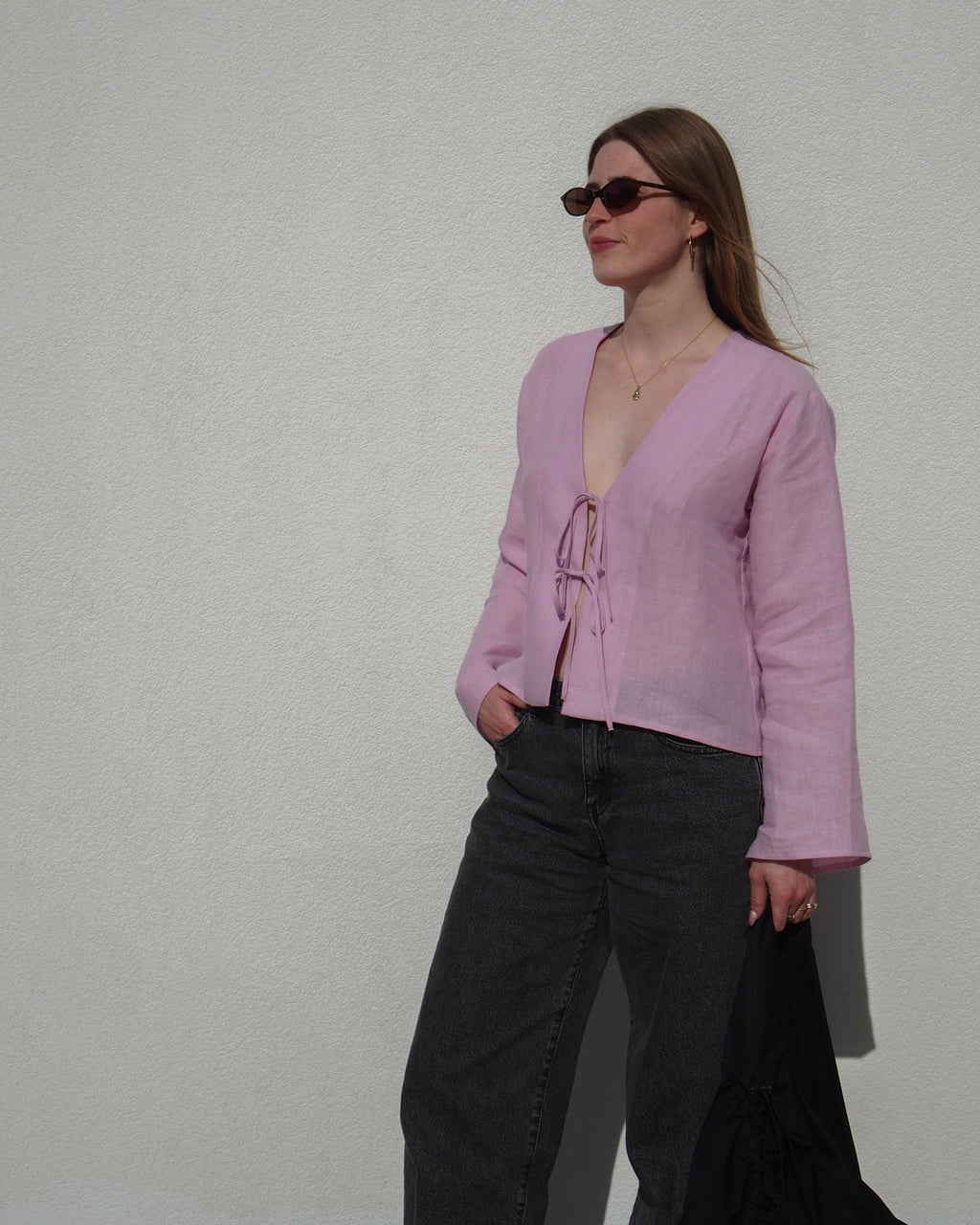 A woman in sunglasses wears the jan-n-june LINEN TIE BLOUSE SKAGEN LIGHT ORCHID with dark jeans, standing against a white wall, holding a black jacket and looking to the side.