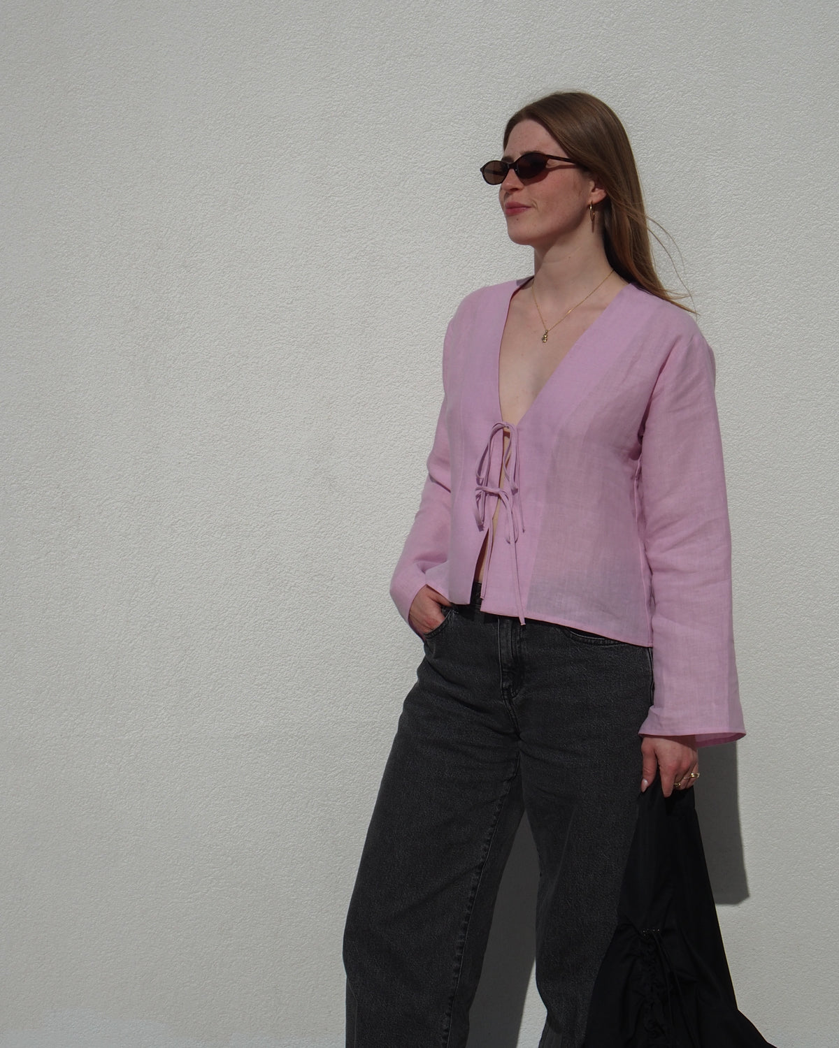 A woman in sunglasses wears the jan-n-june LINEN TIE BLOUSE SKAGEN LIGHT ORCHID with dark jeans, standing against a white wall, holding a black jacket and looking to the side.