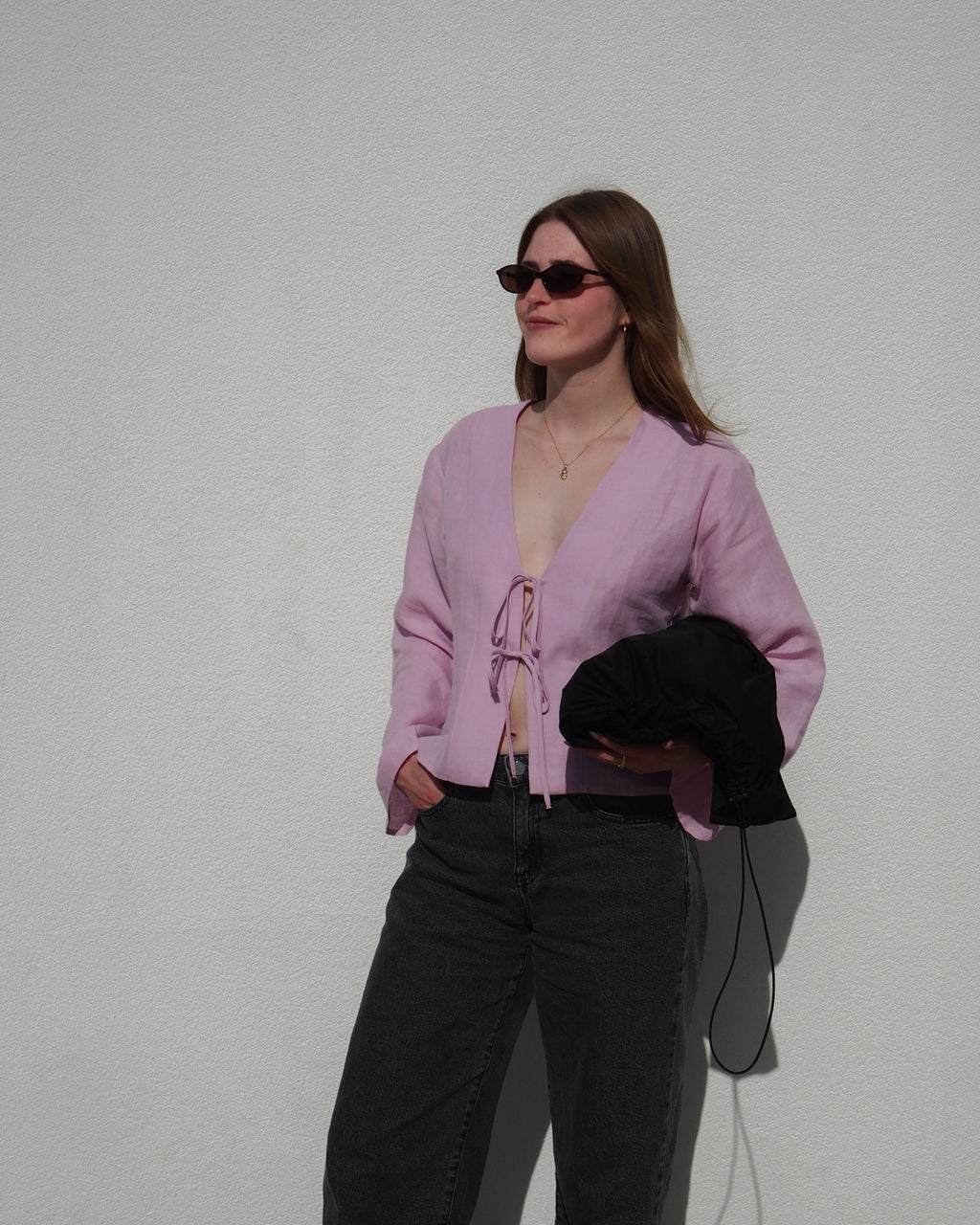 A woman stands against a white wall, wearing jan-n-junes LINEN TIE BLOUSE SKAGEN LIGHT ORCHID in European linen and high-waisted black jeans. She holds a black jacket and smiles gently, looking slightly to the side behind her sunglasses.