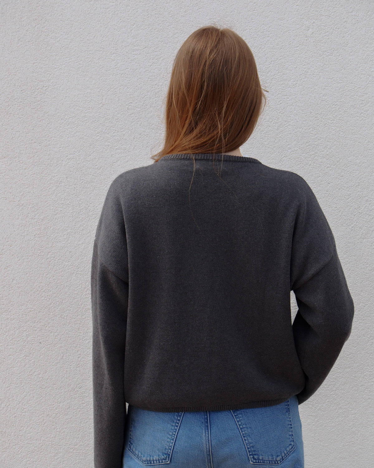 A person with long, light brown hair stands facing a white wall, wearing the jan-n-june LIGHT CARDIGAN ASAS DARK GREY GOTS and light blue jeans. Their back is to the camera, showcasing eco-friendly style.