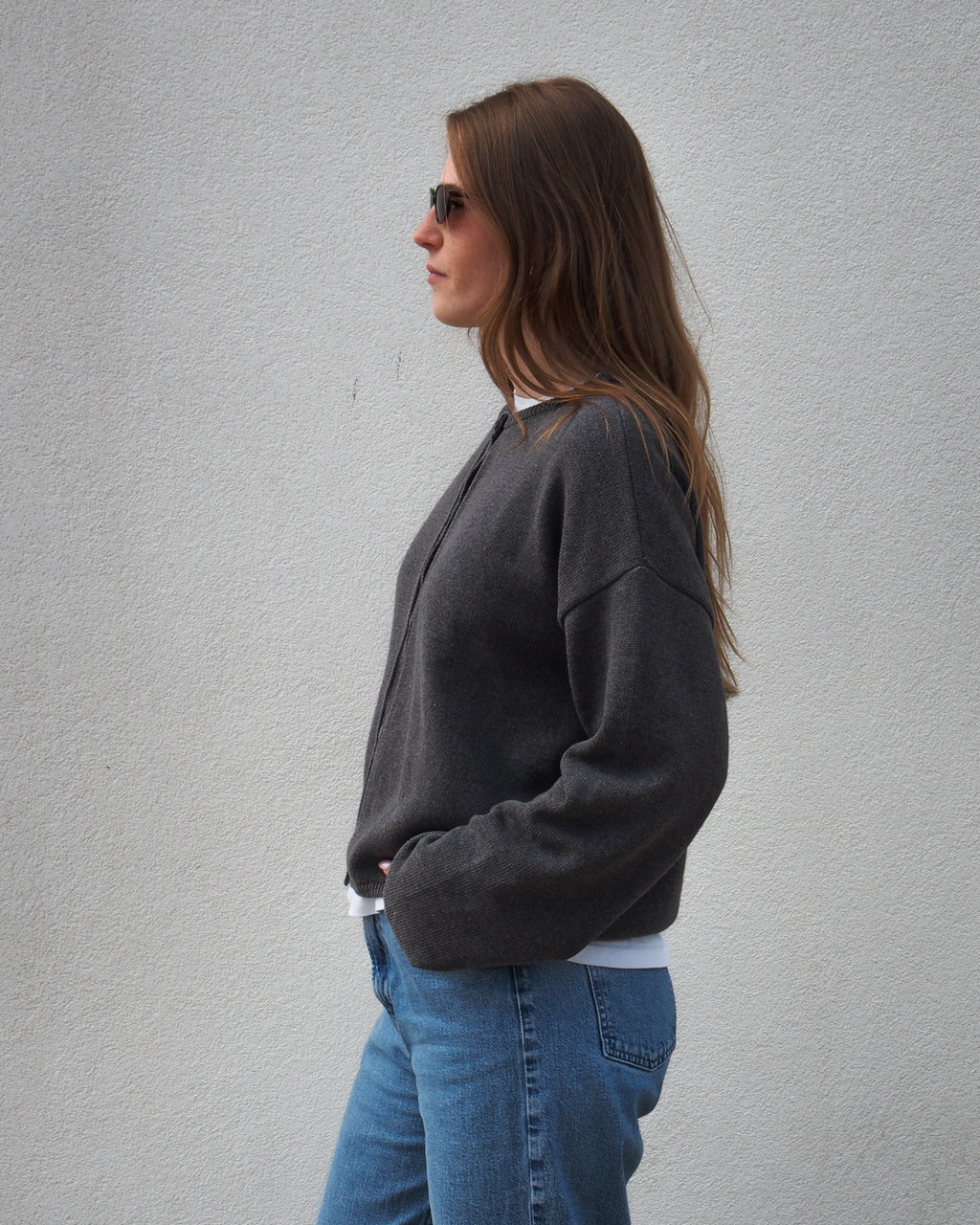 A woman with long brown hair, wearing sunglasses, blue jeans, and the jan-n-june LIGHT CARDIGAN ASAS DARK GREY GOTS, stands in profile against a plain light-colored wall.