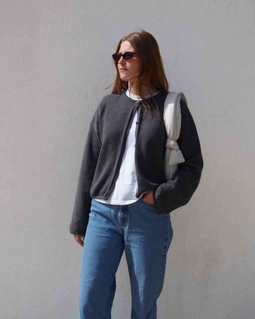 A woman wearing sunglasses and the jan-n-june LIGHT CARDIGAN ASAS DARK GREY GOTS over a white shirt with blue jeans stands against a plain light wall, holding a white padded jacket over one shoulder.