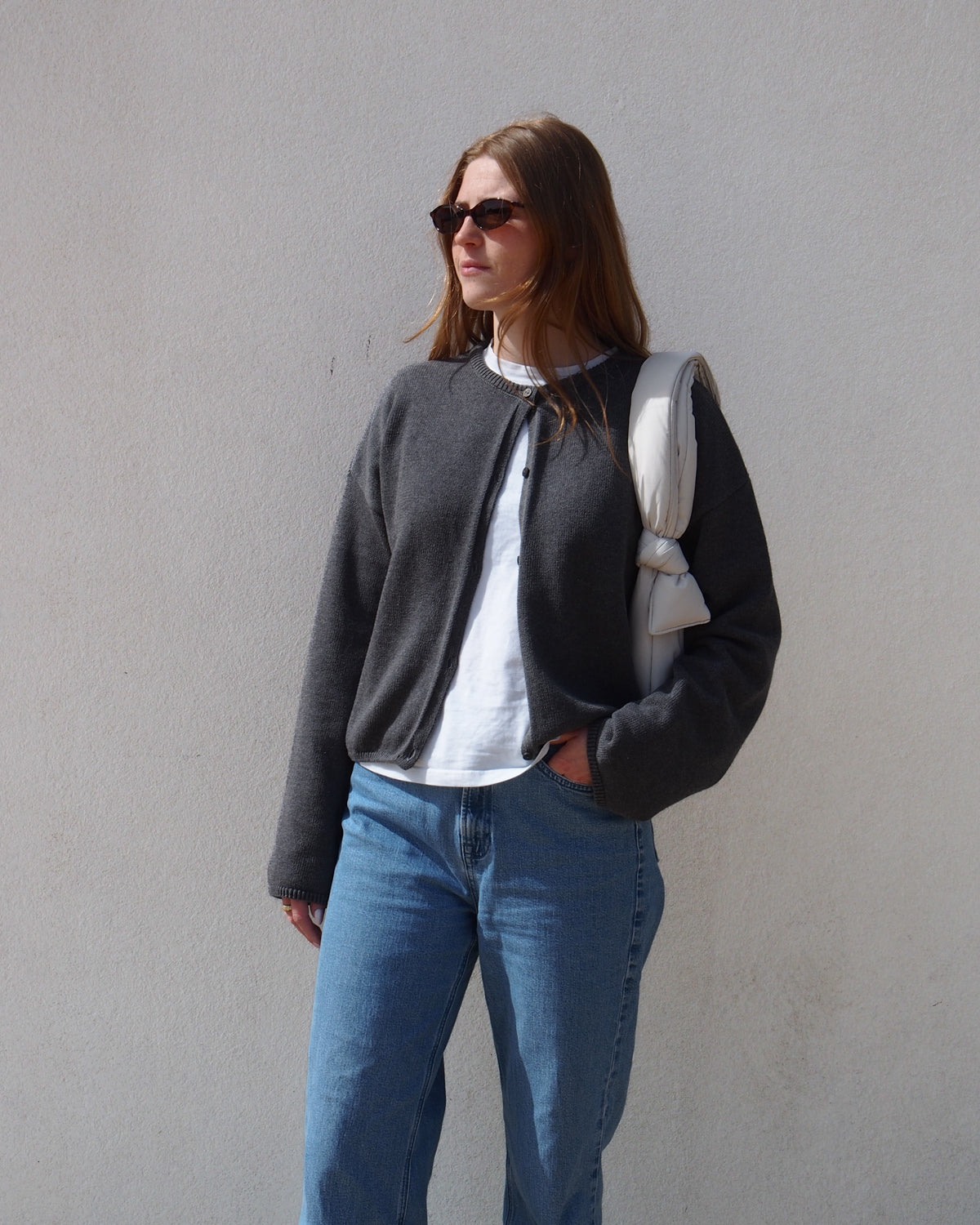 A woman wearing sunglasses and the jan-n-june LIGHT CARDIGAN ASAS DARK GREY GOTS over a white shirt with blue jeans stands against a plain light wall, holding a white padded jacket over one shoulder.