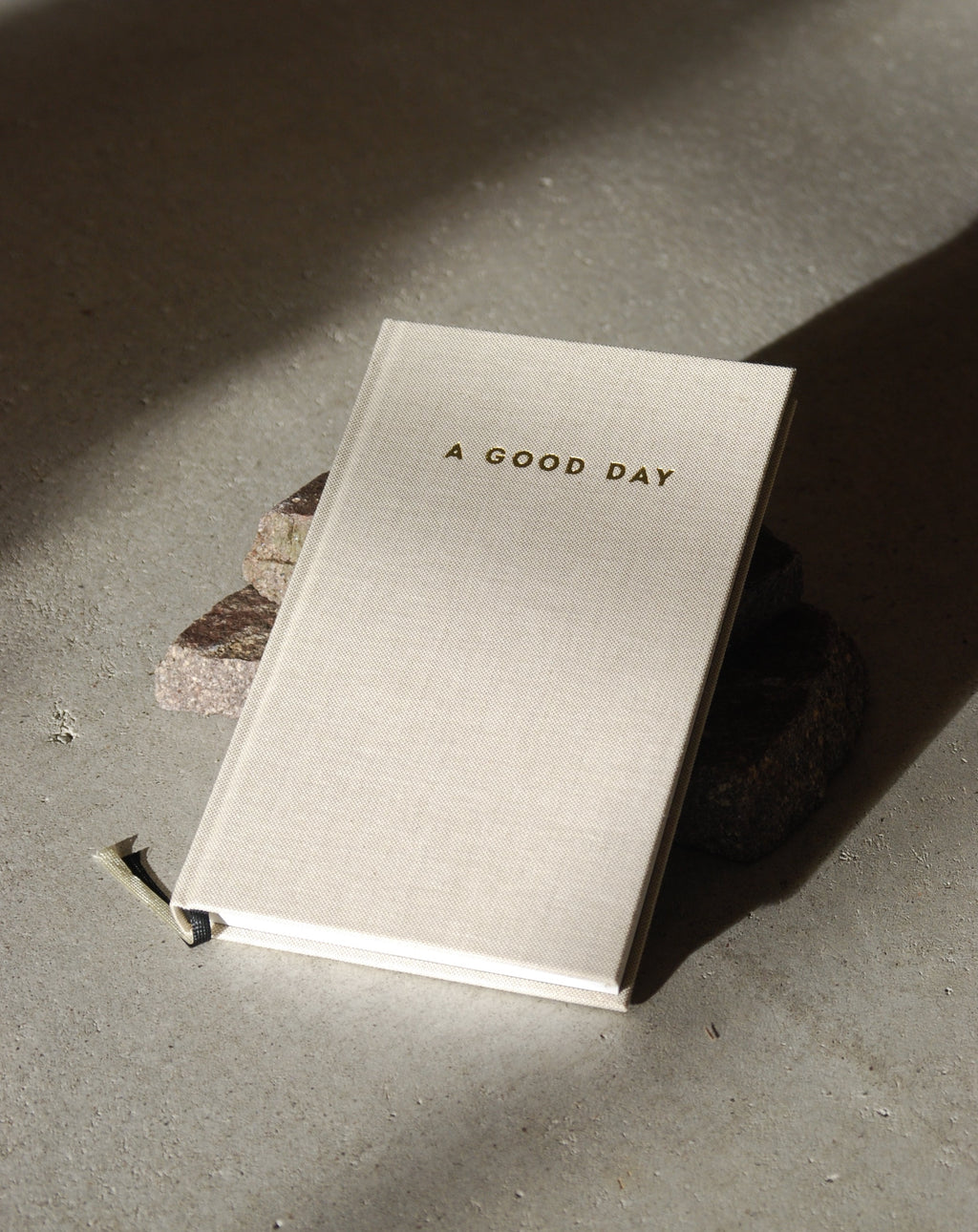 A beige hardcover DIARY A GOOD DAY NATUR by jan-n-june rests on concrete, propped with stones, inviting mindful reflection and self-love as sunlight casts gentle shadows across the scene.