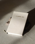 A beige hardcover DIARY A GOOD DAY NATUR by jan-n-june rests on concrete, propped with stones, inviting mindful reflection and self-love as sunlight casts gentle shadows across the scene.