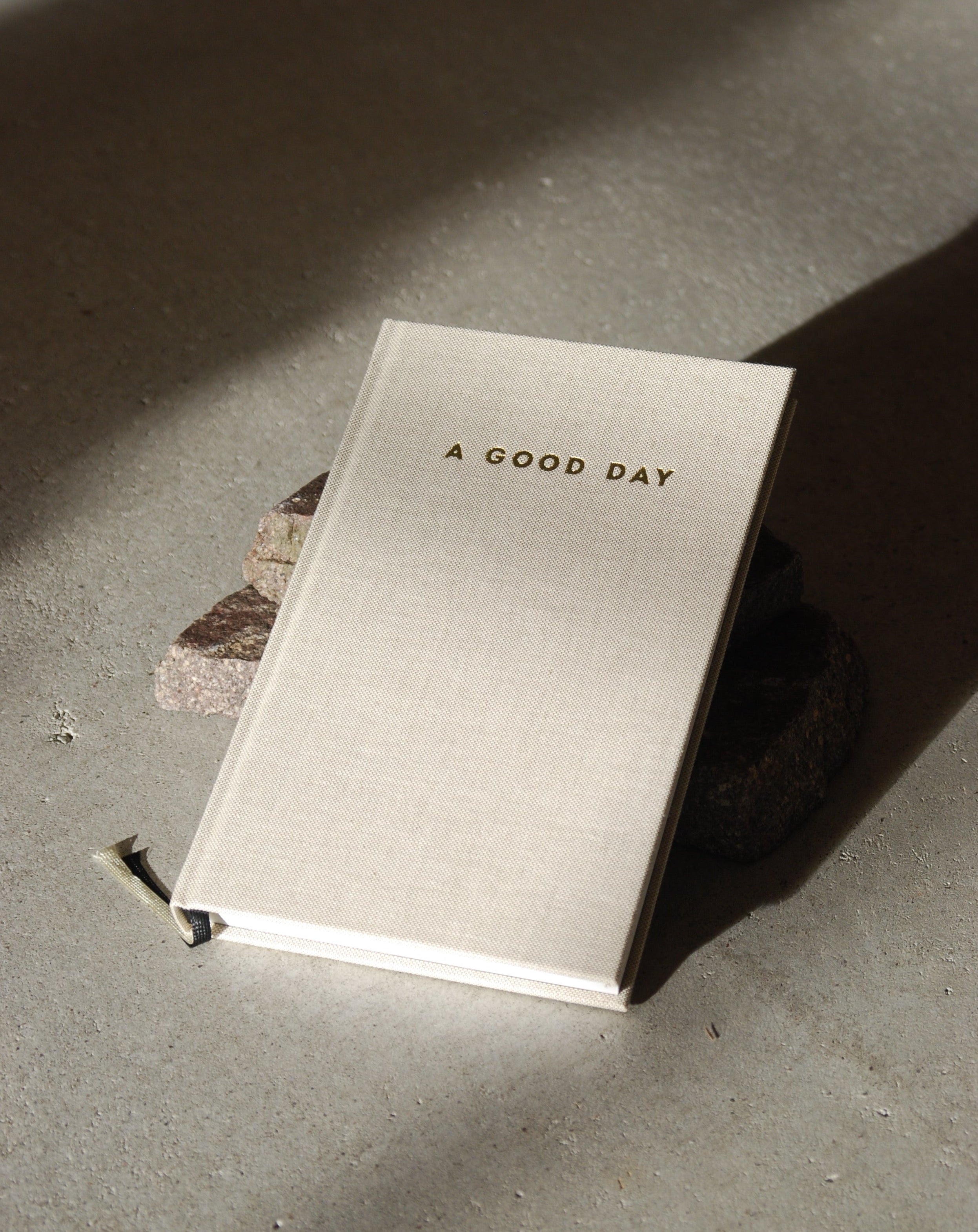 A beige hardcover DIARY A GOOD DAY NATUR by jan-n-june rests on concrete, propped with stones, inviting mindful reflection and self-love as sunlight casts gentle shadows across the scene.