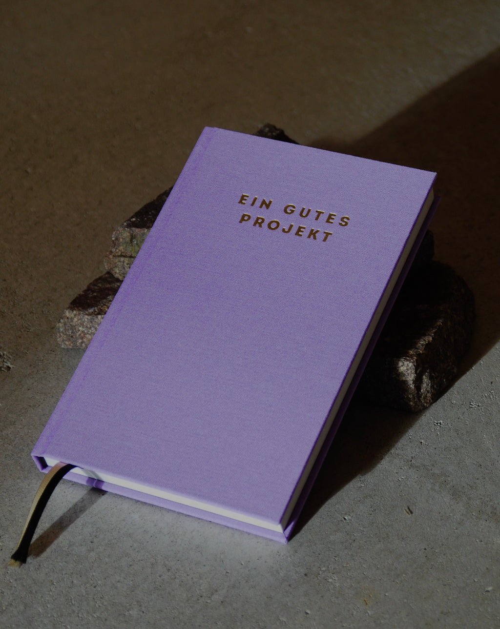 The PLANER EIN GUTES PROJEKT LAVENDER mindful success journal by jan-n-june, featuring a lavender hardcover with gold lettering and a black ribbon bookmark, is placed on dark stones on a gray surface.