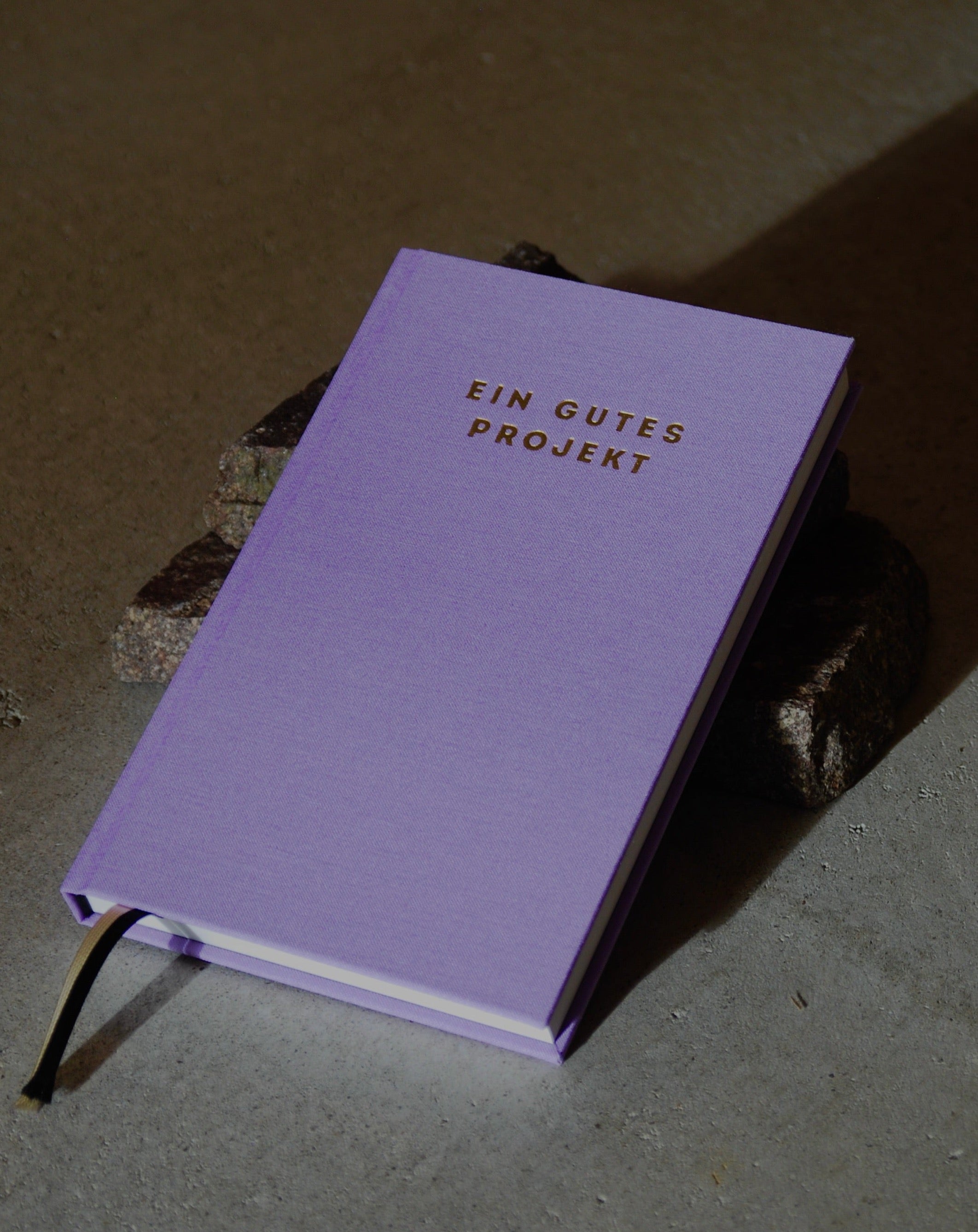 The PLANER EIN GUTES PROJEKT LAVENDER mindful success journal by jan-n-june, featuring a lavender hardcover with gold lettering and a black ribbon bookmark, is placed on dark stones on a gray surface.