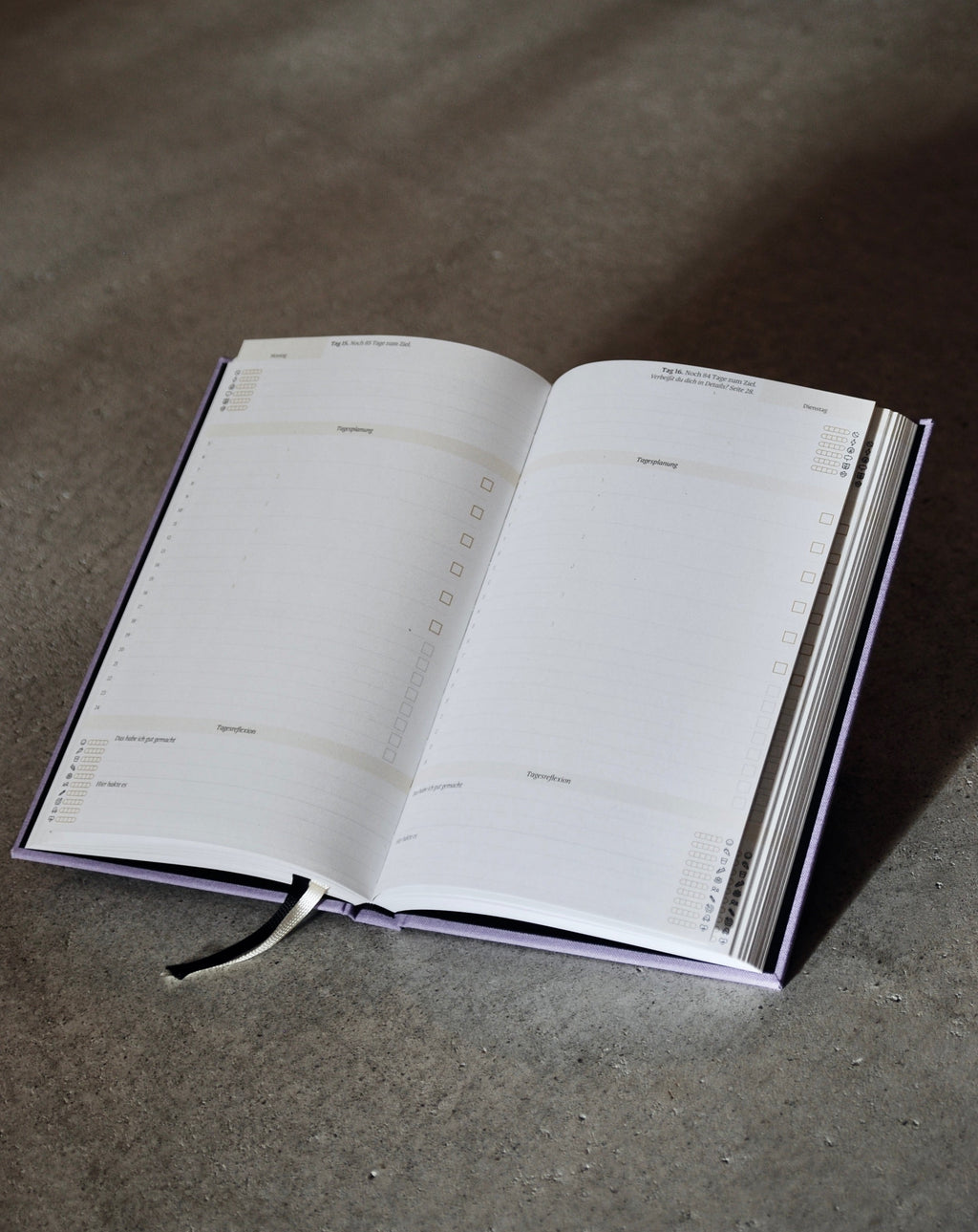 The PLANER EIN GUTES PROJEKT LAVENDER by jan-n-june is an open mindful success journal with blank, sustainably made pages labeled by weekdays and sections for notes or tasks, plus checkboxes and a ribbon bookmark, on a gray surface.