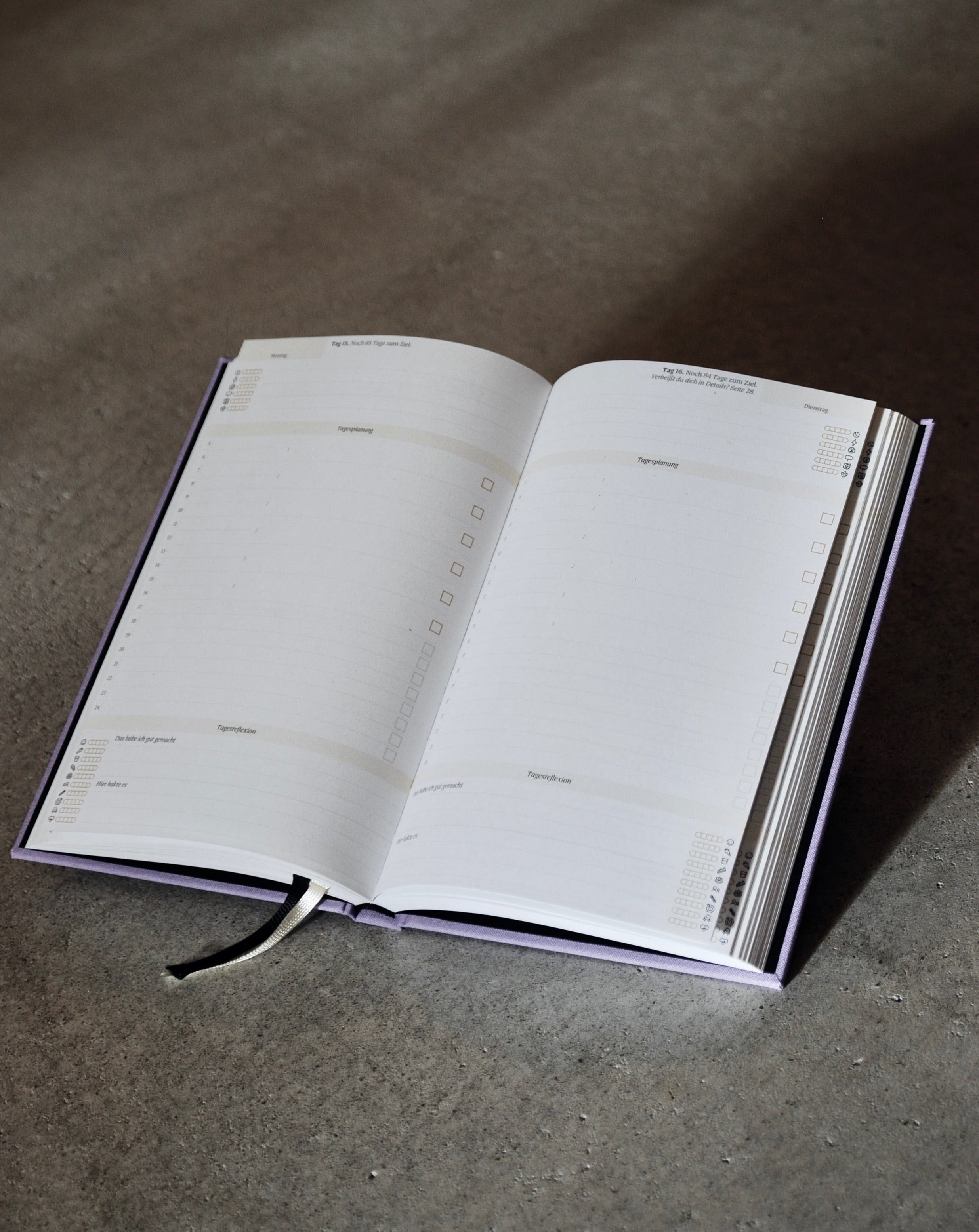 The PLANER EIN GUTES PROJEKT LAVENDER by jan-n-june is an open mindful success journal with blank, sustainably made pages labeled by weekdays and sections for notes or tasks, plus checkboxes and a ribbon bookmark, on a gray surface.