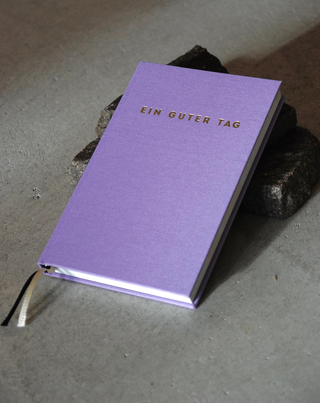 The jan-n-june TAGEBUCH EIN GUTER TAG LAVENDER notebook features gold lettering on a lavender cover—resting elegantly on dark stones, it’s an ideal companion for mindfulness and self-love journaling.