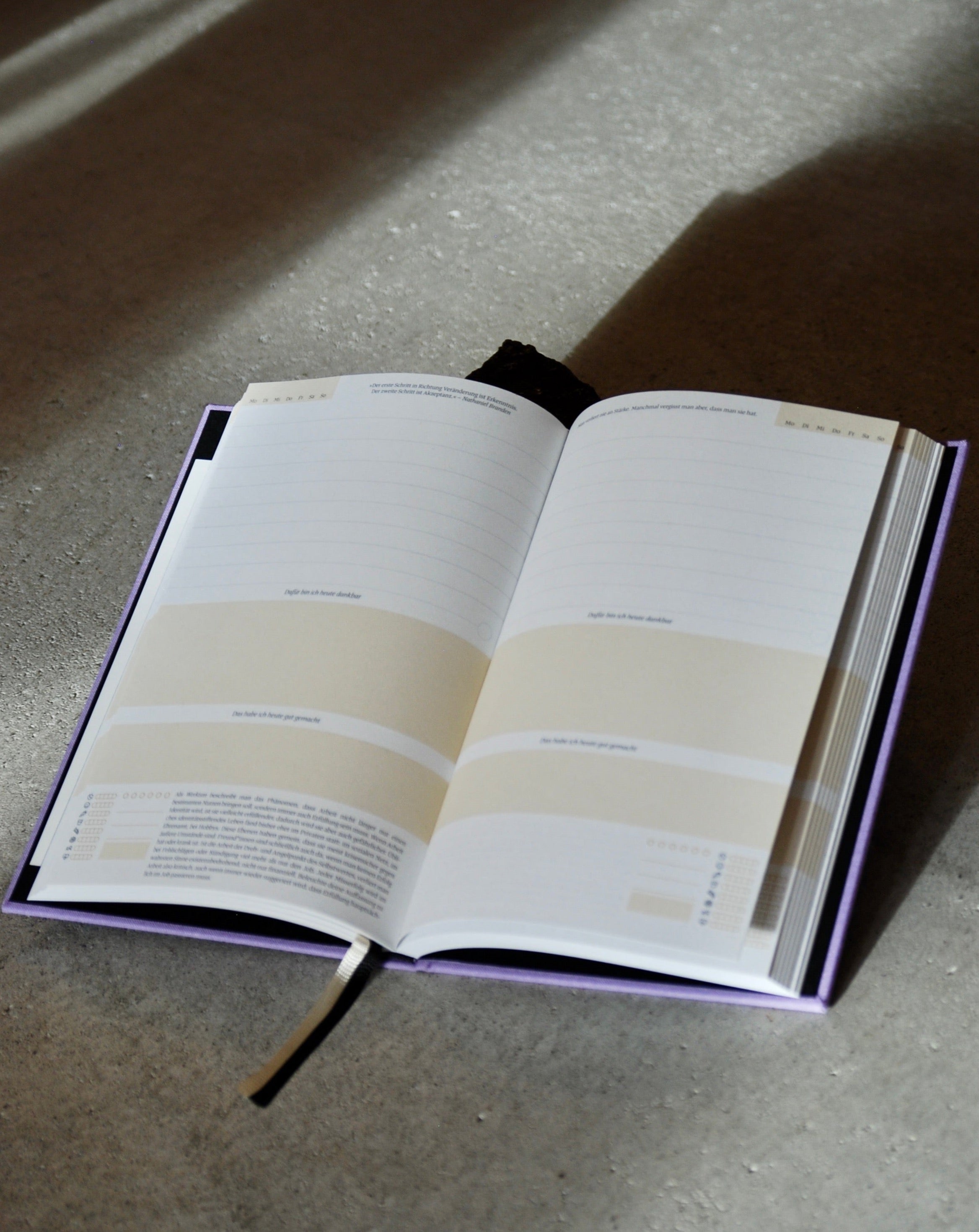The jan-n-june TAGEBUCH EIN GUTER TAG LAVENDER, open on a gray surface with a black ribbon and off-white fabric bookmark, invites mindfulness and reflection with its lined and shaded pages partly bathed in shadow.