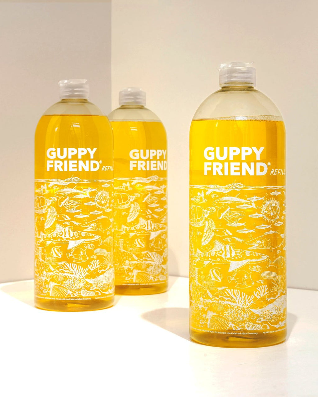 Three clear plastic bottles of GUPPYFRIEND LAUNDRY DETERGENT by jan-n-june, filled with yellow liquid and decorated with white sea life illustrations, stand on a white surface. These detergent bottles are microplastic-free.