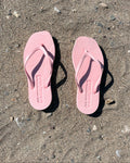 The jan-n-june SLEEPERS TAPERED FLIP FLOPS in Pink Sorbet, crafted from natural rubber, rest side by side on sandy ground with scattered pebbles and stones, casting clear shadows in the sunlight.