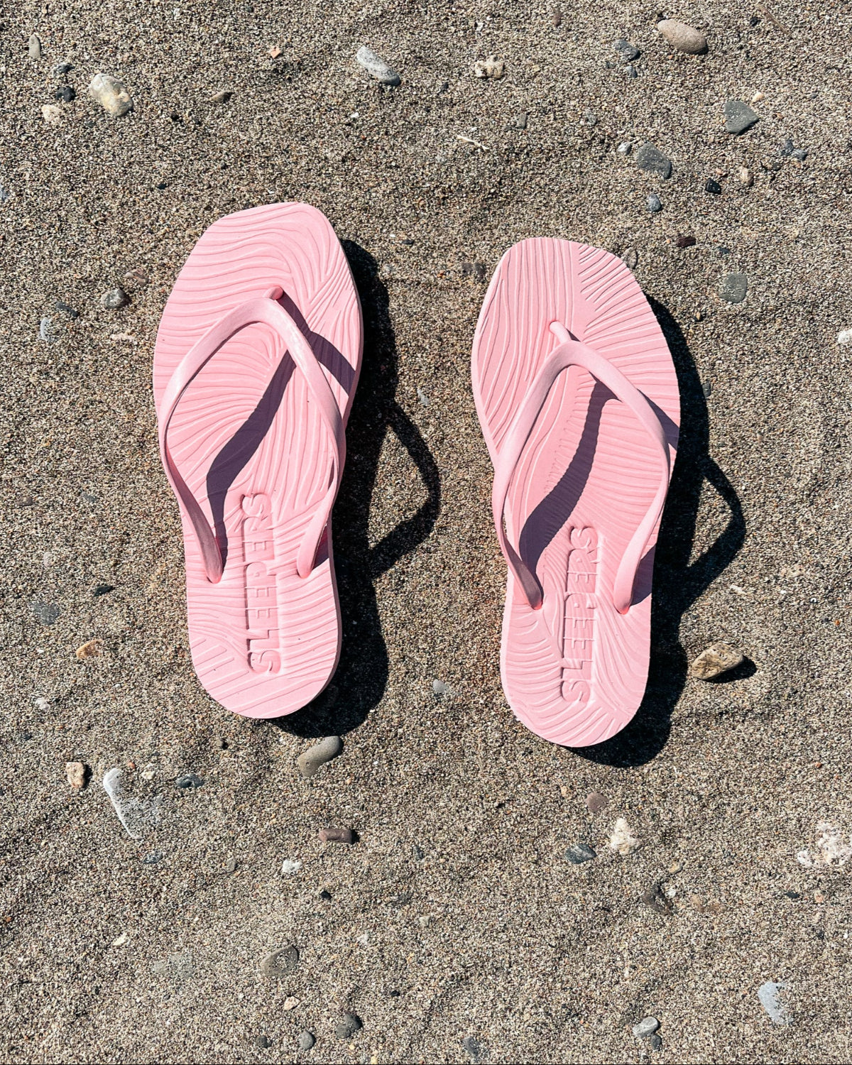 The jan-n-june SLEEPERS TAPERED FLIP FLOPS in Pink Sorbet, crafted from natural rubber, rest side by side on sandy ground with scattered pebbles and stones, casting clear shadows in the sunlight.