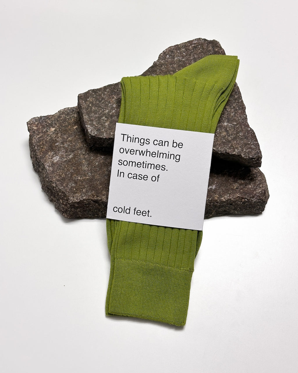 A pair of jan-n-june SOCKS MOSSY PEAR made from organic cotton rests on two rough stones. A card on top reads, Things can be overwhelming sometimes. In case of cold feet. The background is plain white.