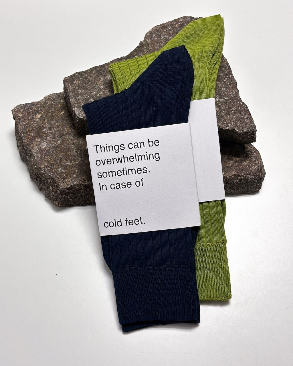 Two pairs of jan-n-june SOCKS MOSSY PEAR, one navy and one olive green, made from organic cotton, rest on stacked rocks. Their paper label says: Things can be overwhelming sometimes. In case of cold feet.