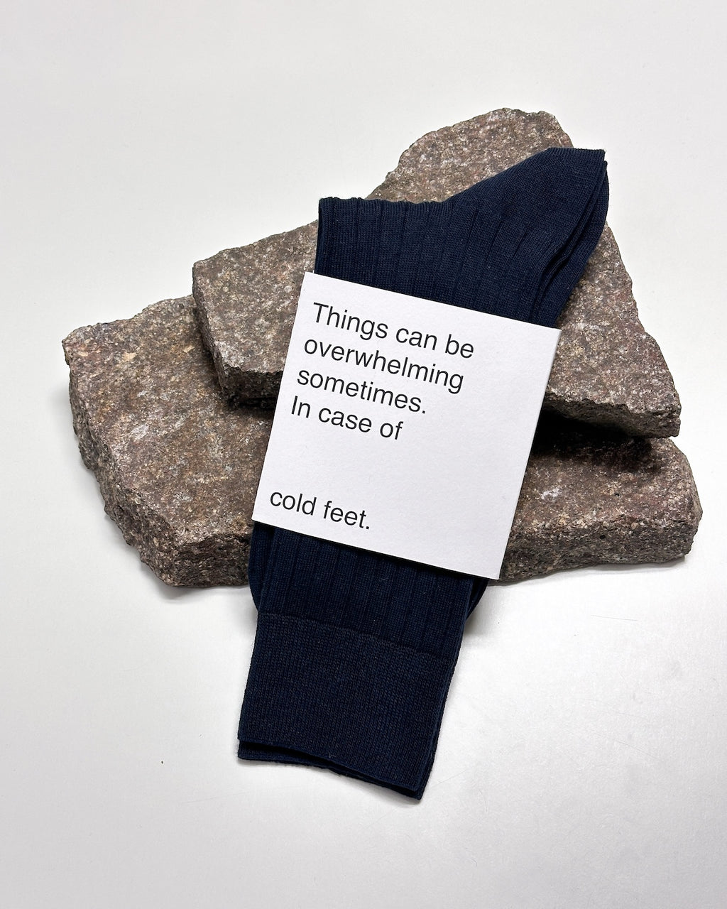 A pair of dark blue SOCKS NIGHTFALL by jan-n-june, made from organic cotton, rests on stacked rough stones. The label reads: Things can be overwhelming sometimes. In case of cold feet.