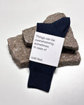 A pair of dark blue SOCKS NIGHTFALL by jan-n-june, made from organic cotton, rests on stacked rough stones. The label reads: Things can be overwhelming sometimes. In case of cold feet.